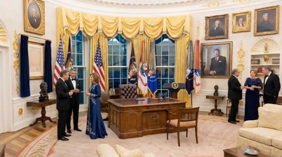 People in formal attire gather in the Oval Office, engaging in conversation and admiring decor
