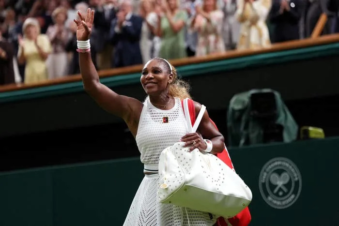 Athlete in a tennis outfit waves to the crowd on a sports court, carrying a large bag